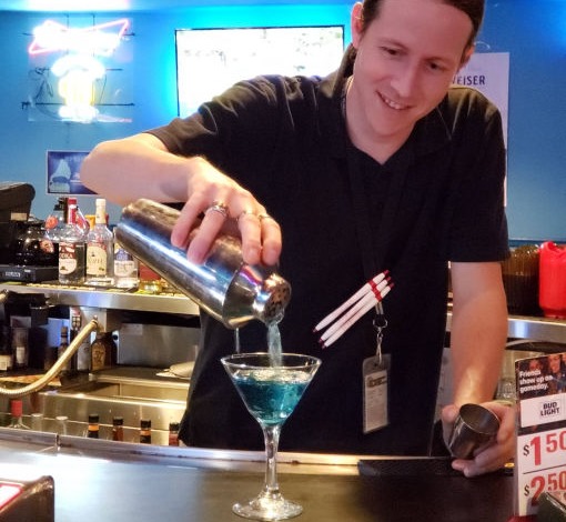 A bartender in a black shirt is smiling as he pours a vibrant blue cocktail into a martini glass from a metal shaker. The bar area has various bottles displayed in the background, with a neon sign and a few visible drink specials on the counter.