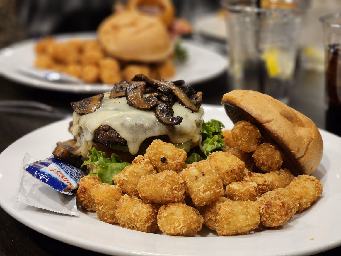 "A plate of a juicy burger topped with melted cheese, grilled mushrooms, and fresh lettuce, served with crispy golden tater tots and a side of condiment packet."
