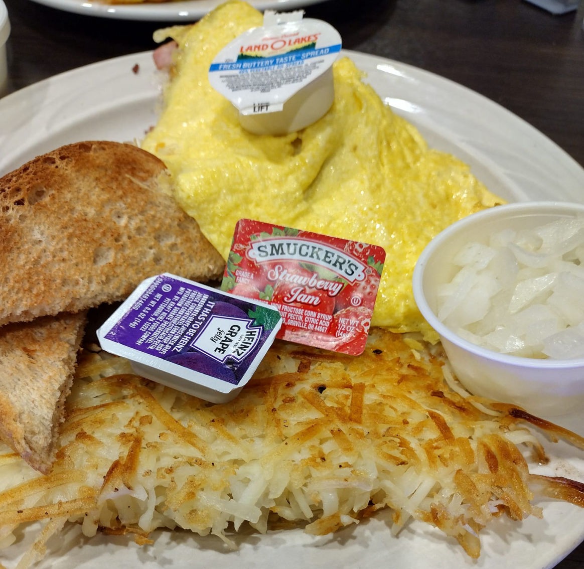 A plate of breakfast with a fluffy omelette, crispy hash browns, two slices of toasted bread, and assorted condiments including grape jelly, strawberry jam, butter spread, and onions on the side.