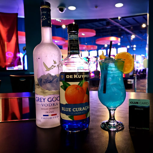 A vibrant blue cocktail with an orange garnish sits on a bar counter next to bottles of Grey Goose vodka and De Kuyper Blue Curacao, with a High Winds Casino Breeze Club Card in the foreground. The casino slot machines are softly blurred in the background.