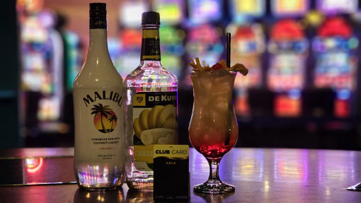 "A tropical cocktail with a cherry and fruit garnish sits in front of a blurry backdrop of slot machines. The drink is accompanied by bottles of Malibu coconut rum and De Kuyper banana liqueur. A High Winds Casino Gale Club Card rests on the bar next to the bottles."
