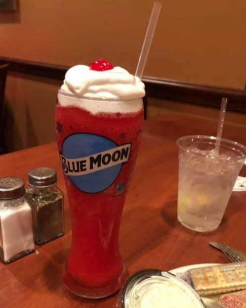 A close-up of a Blue Moon glass filled with a vibrant red frozen drink topped with whipped cream and a cherry. The glass has the Blue Moon logo on it. In the background, there is a clear cup of water and a plate with some food items.
