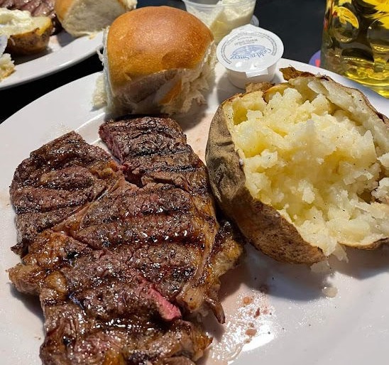 A plate of grilled steak with a baked potato, butter, and two rolls on the side. The baked potato is partially scooped out with a soft interior, ready to be topped.